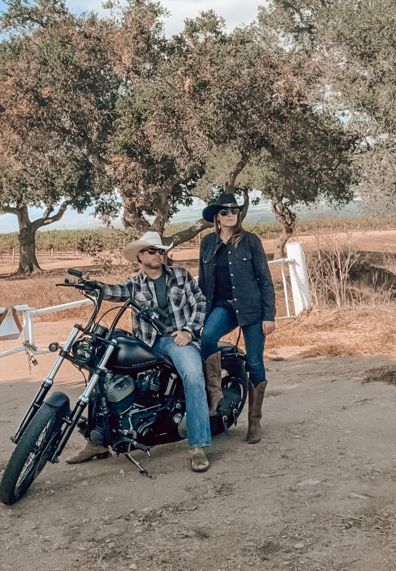 Two people sitting on a motorcycle in a rural setting with trees and open space.