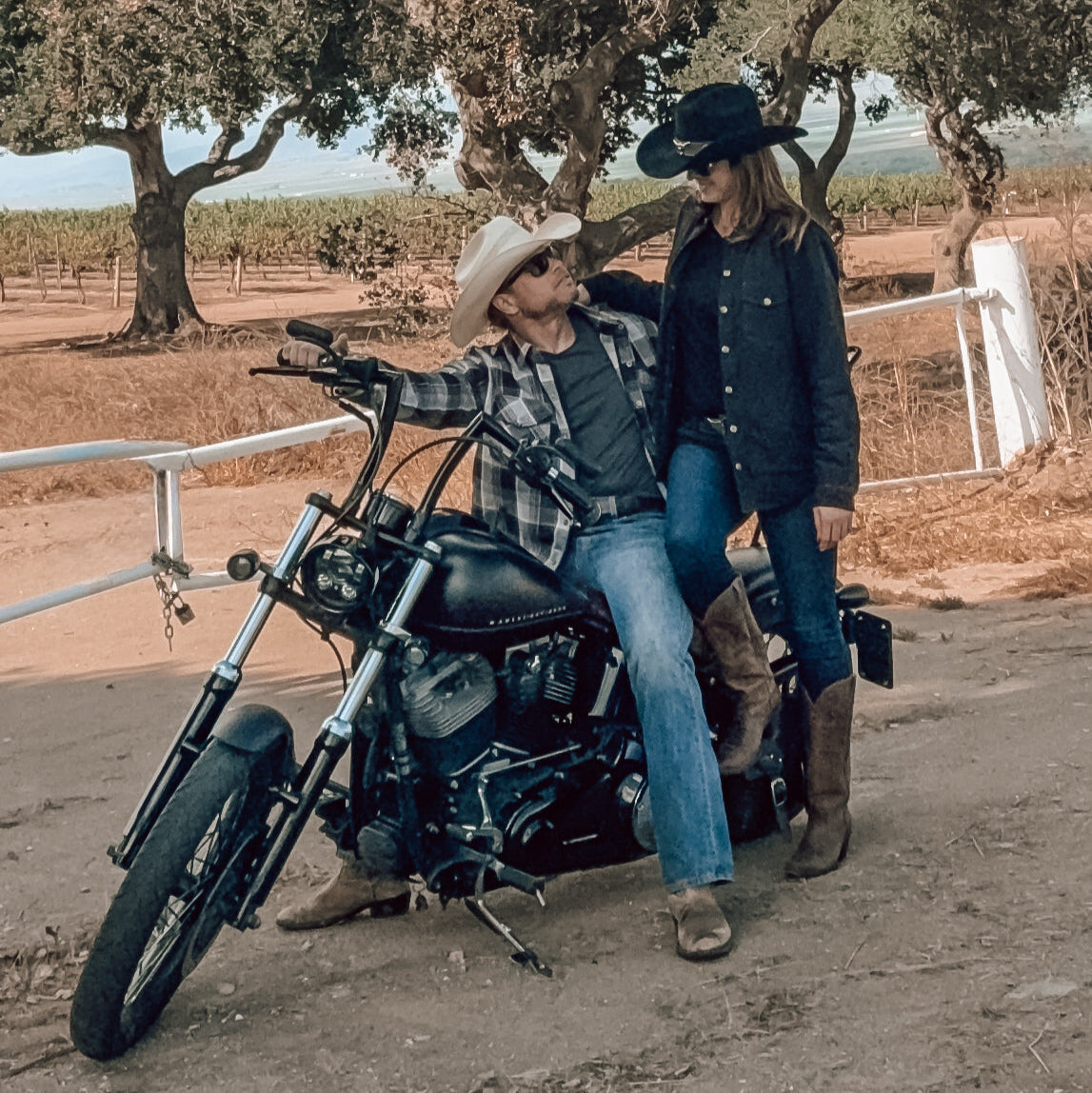 Two people on a motorcycle in a rural setting with trees and open space.